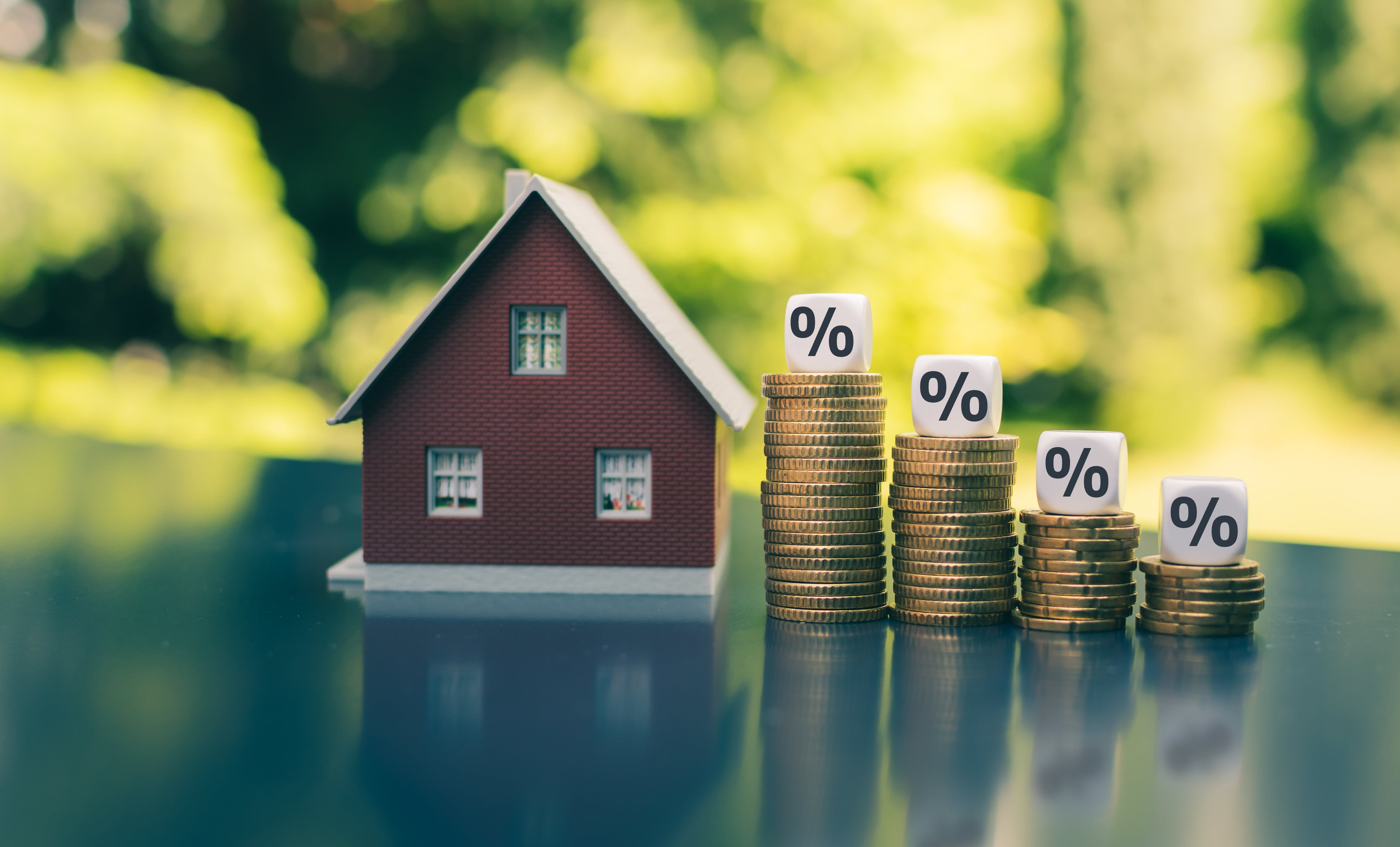 A small red house model beside stacks of gold coins with percentage symbols on top. A small red house model beside stacks of gold coins with percentage symbols on top.